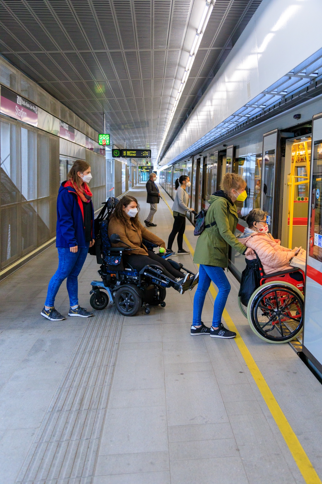 Im Vordergrund steigen zwei weibliche Fahrgäste im Rollstuhl mit Hilfe ihrer Assistentinnen in eine U-Bahn ein. Im Hintergrund ist eine Dame mit Kinderwagen und ein blinder Fahrgast mit Langstock beim Einstieg zu sehen. Alle tragen FFP2 Masken.